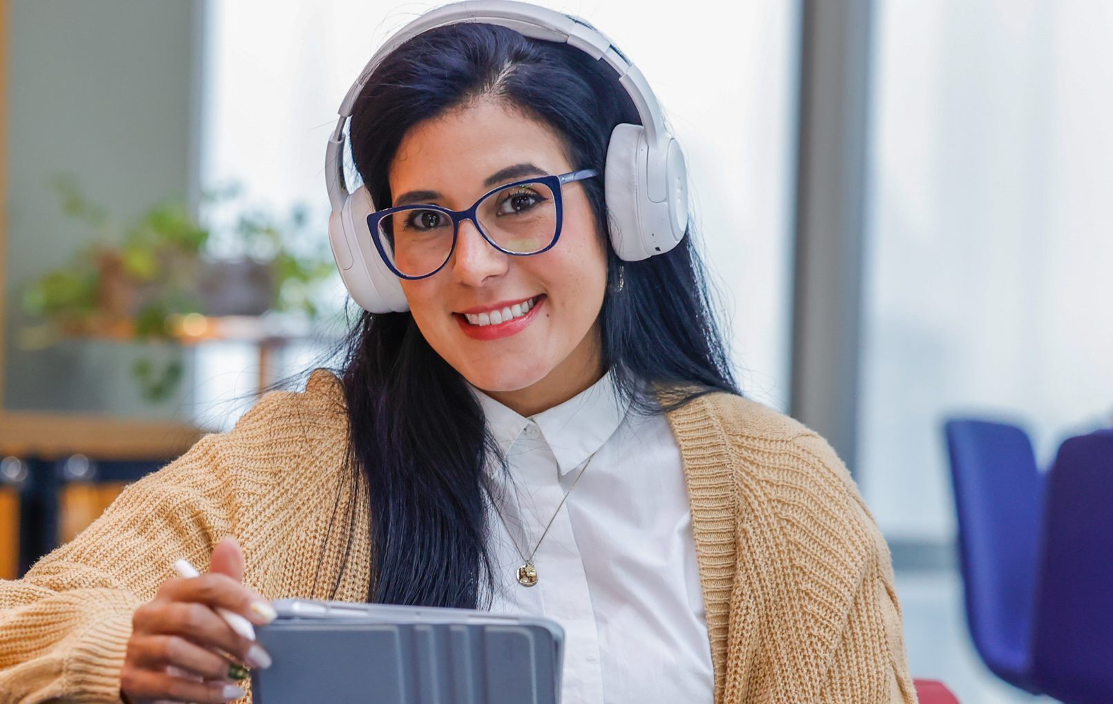 Mulher sorrindo, de óculos e fones de ouvido, segurando um tablet e uma caneta digital em um ambiente de estudo.
